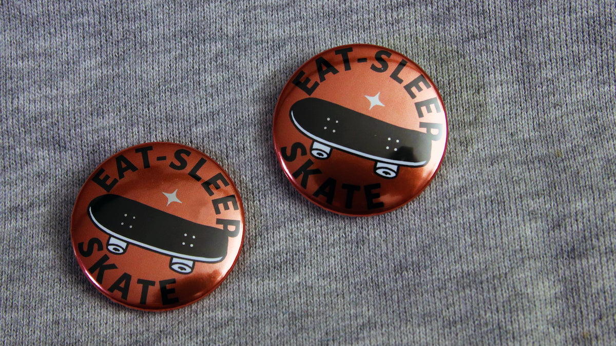 32mm (1.25 inch) silver Eat Sleep Skate logo pinned to a grey sweater