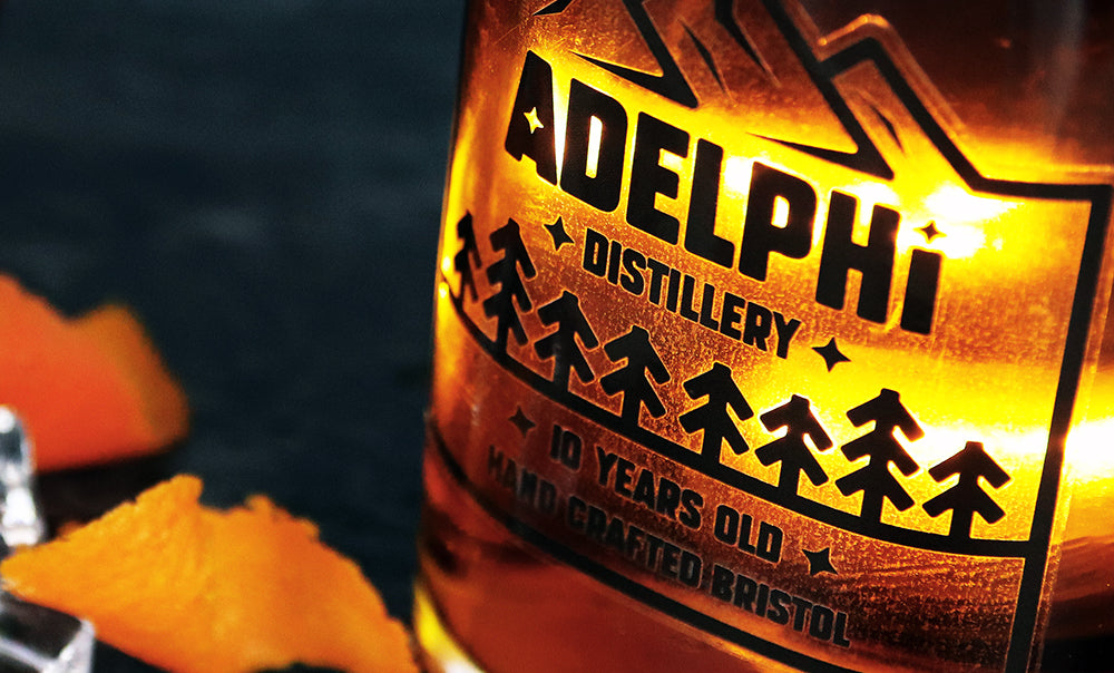 Clear die cut sticker applied to a whiskey bottle next to a filled jar, orange peel, ice cubes and cards