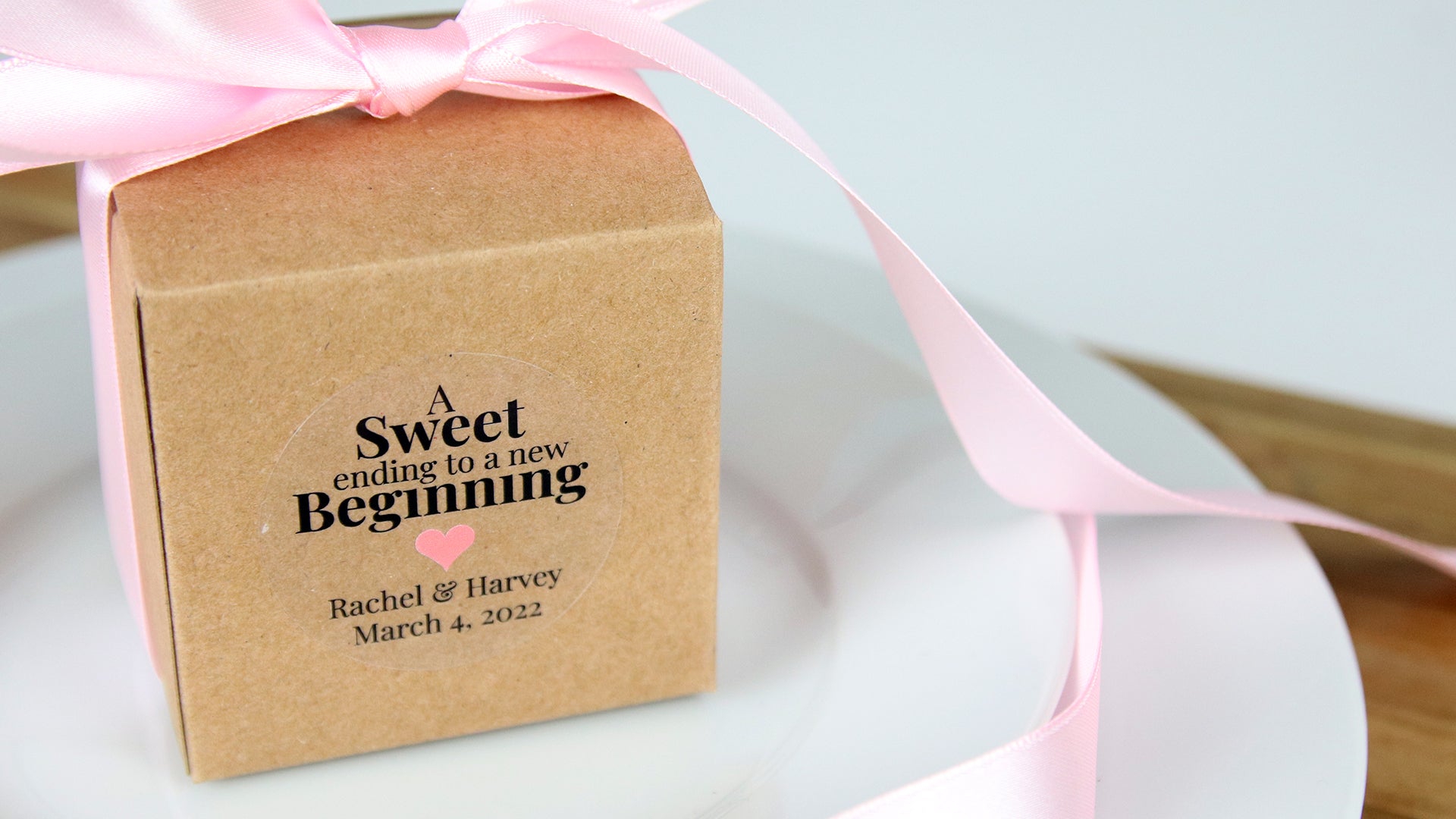 Round clear sticker applied to a cardboard box with a pink bow containing a wedding gift for guest on a white plate
