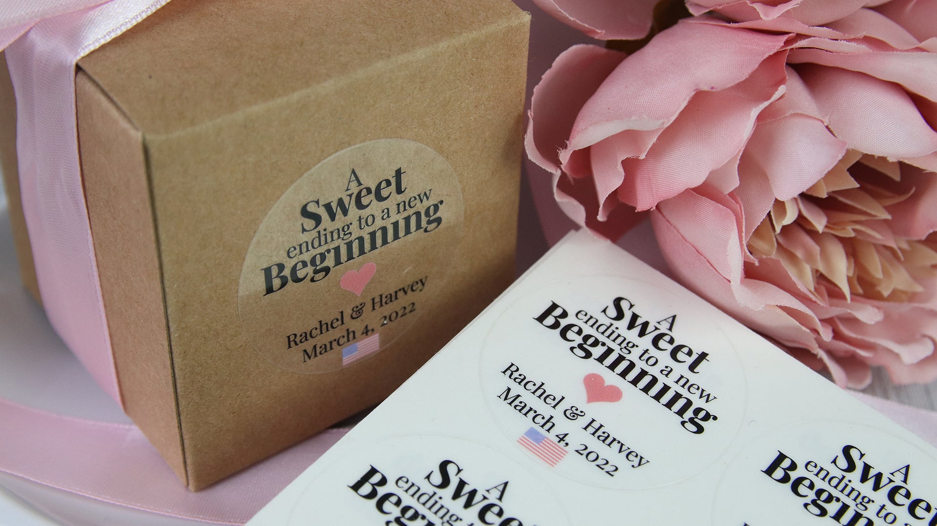 Round clear sticker from a sheet on a box with a pink bow containing a wedding gift for guest on a white plate next to sticker sheets