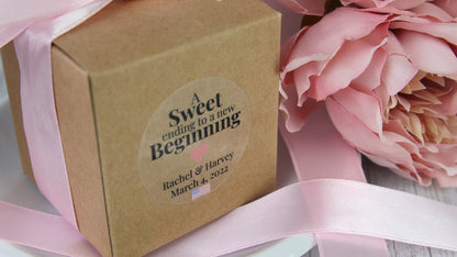 Round clear sticker applied to a cardboard box with a pink bow containing a wedding gift for guest on a white plate